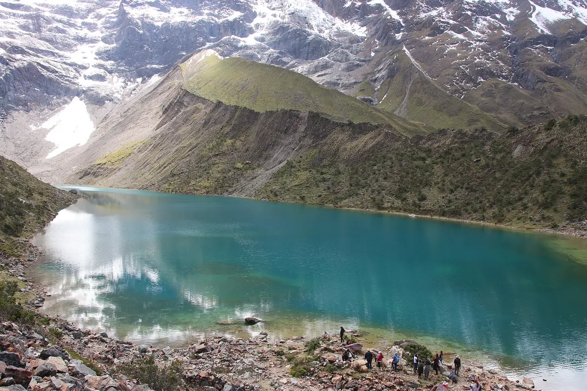 Lago Humantay com água turquesa sob um glaciar suspenso e encostas íngremes, com algumas pessoas na margem pedregosa